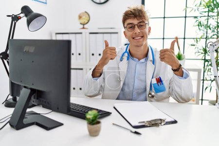 Téléchargez les photos : Young caucasian doctor man working at the clinic success sign doing positive gesture with hand, thumbs up smiling and happy. cheerful expression and winner gesture. - en image libre de droit