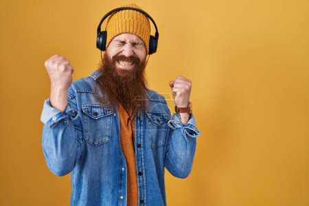 Photo for Caucasian man with long beard listening to music using headphones very happy and excited doing winner gesture with arms raised, smiling and screaming for success. celebration concept. - Royalty Free Image