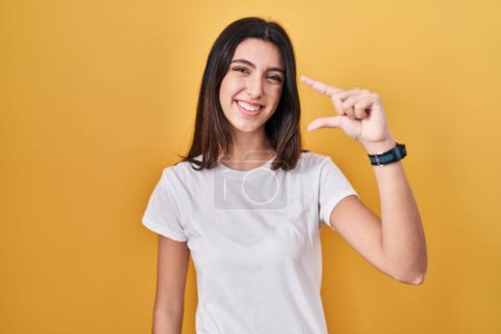 Téléchargez les photos : Young beautiful woman standing over yellow background smiling and confident gesturing with hand doing small size sign with fingers looking and the camera. measure concept. - en image libre de droit