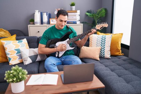 Photo for Young hispanic man having online electrical guitar class sitting on sofa at home - Royalty Free Image