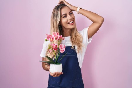 Foto de Joven mujer rubia vistiendo delantal jardinero sosteniendo planta sonriente seguro de tocar el cabello con la mano hacia arriba gesto, posando atractivo y de moda - Imagen libre de derechos