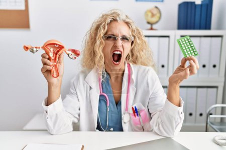 Foto de Médico de mediana edad mujer sosteniendo píldoras anticonceptivas enojado y loco gritando frustrado y furioso, gritando con ira. rabia y concepto agresivo. - Imagen libre de derechos