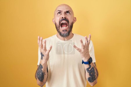 Téléchargez les photos : Homme hispanique avec des tatouages debout sur fond jaune fou et fou crier et crier avec une expression agressive et les bras levés. concept de frustration. - en image libre de droit