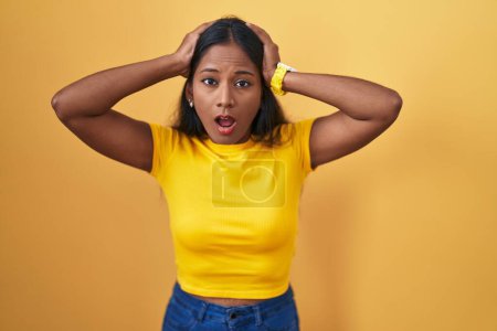 Photo for Young indian woman standing over yellow background crazy and scared with hands on head, afraid and surprised of shock with open mouth - Royalty Free Image