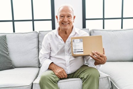 Foto de Hombre mayor sosteniendo paquete de entrega en casa con aspecto positivo y feliz de pie y sonriendo con una sonrisa confiada mostrando los dientes - Imagen libre de derechos