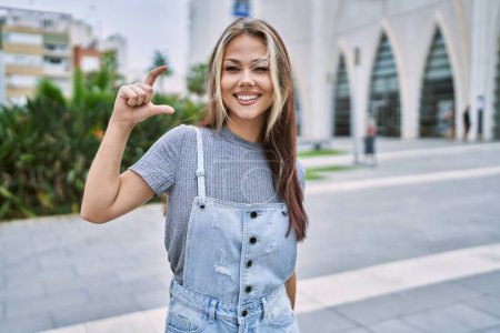 Photo for Young caucasian woman outdoors smiling and confident gesturing with hand doing small size sign with fingers looking and the camera. measure concept. - Royalty Free Image