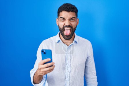 Téléchargez les photos : Homme hispanique avec barbe en utilisant le message de dactylographie smartphone langue collante heureux avec une expression drôle. concept d'émotion. - en image libre de droit