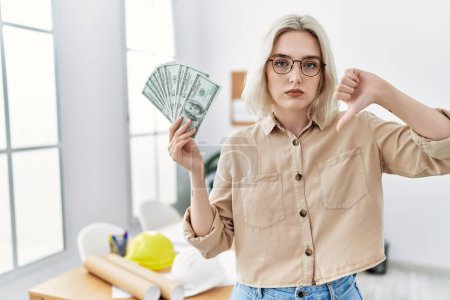 Photo for Young beautiful caucasian woman at construction office holding money with angry face, negative sign showing dislike with thumbs down, rejection concept - Royalty Free Image