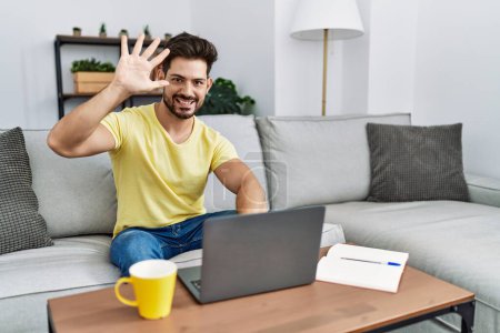 Photo for Young man with beard using laptop at home showing and pointing up with fingers number five while smiling confident and happy. - Royalty Free Image
