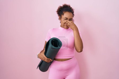 Foto de Young hispanic woman with curly hair holding yoga mat over pink background smelling something stinky and disgusting, intolerable smell, holding breath with fingers on nose. bad smell - Imagen libre de derechos