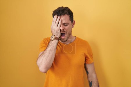 Photo for Young hispanic man standing over yellow background yawning tired covering half face, eye and mouth with hand. face hurts in pain. - Royalty Free Image