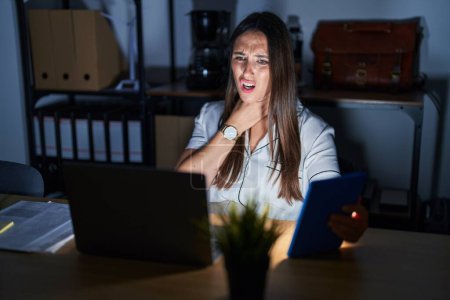 Foto de Young brunette woman working at the office at night touching painful neck, sore throat for flu, clod and infection - Imagen libre de derechos
