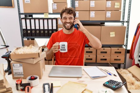 Foto de Joven hombre hispano usando smartphone y bebiendo una taza de café en la oficina sonriendo y riendo en voz alta porque gracioso chiste loco. - Imagen libre de derechos