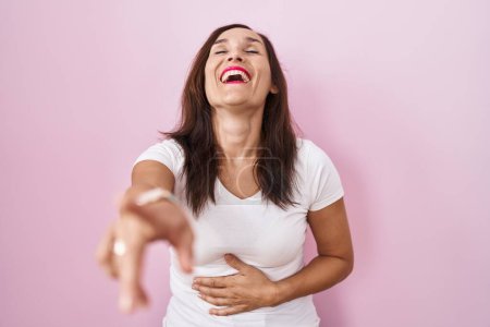 Foto de Mujer morena de mediana edad de pie sobre fondo rosa riéndose de ti, señalando con el dedo a la cámara con la mano sobre el cuerpo, expresión de vergüenza - Imagen libre de derechos