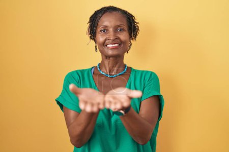 Foto de Mujer africana con rastas de pie sobre fondo amarillo sonriendo con las palmas de las manos juntas recibiendo o dando gesto. retención y protección - Imagen libre de derechos