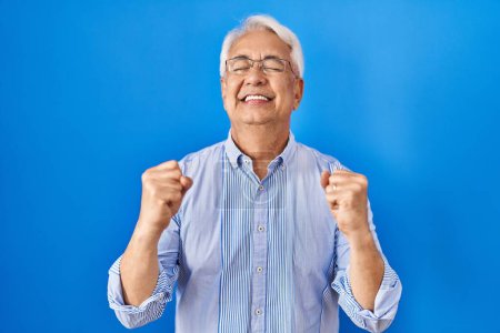 Foto de Hombre mayor hispano con gafas emocionadas por el éxito con los brazos levantados y los ojos cerrados celebrando la victoria sonriendo. concepto ganador. - Imagen libre de derechos
