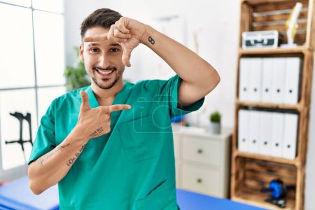 Photo for Young physiotherapist man working at pain recovery clinic smiling making frame with hands and fingers with happy face. creativity and photography concept. - Royalty Free Image