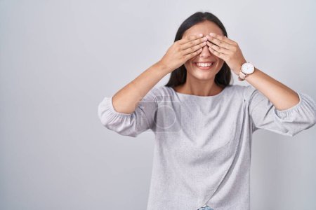 Foto de Young hispanic woman standing over white background covering eyes with hands smiling cheerful and funny. blind concept. - Imagen libre de derechos
