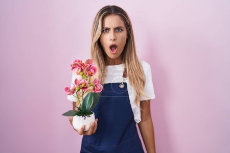 Photo for Young blonde woman wearing gardener apron holding plant in shock face, looking skeptical and sarcastic, surprised with open mouth - Royalty Free Image
