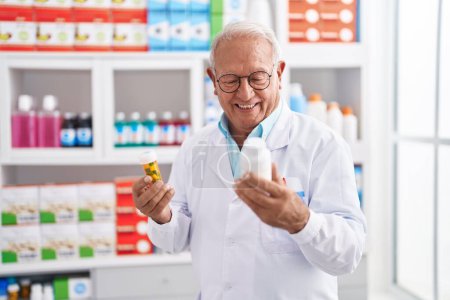 Téléchargez les photos : Homme âgé aux cheveux gris pharmacien souriant confiant tenant des bouteilles de pilules à la pharmacie - en image libre de droit