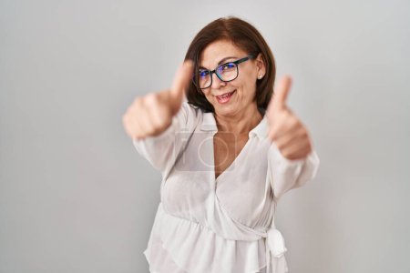 Foto de Mujer hispana de mediana edad de pie sobre un fondo aislado aprobando hacer un gesto positivo con la mano, pulgares arriba sonriendo y feliz por el éxito. gesto ganador. - Imagen libre de derechos