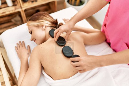 Photo for Young caucasian woman lying on table having back massage using hot stones at beauty salon - Royalty Free Image