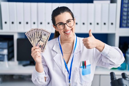 Téléchargez les photos : Jeune femme brune travaillant au laboratoire scientifique tenant de l'argent souriant heureux et positif, pouce levé faire excellent et signe d'approbation - en image libre de droit