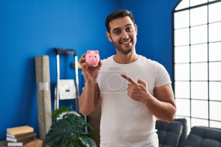 Téléchargez les photos : Jeune homme hispanique avec barbe tenant tirelire à la nouvelle maison souriant heureux pointant avec la main et le doigt - en image libre de droit
