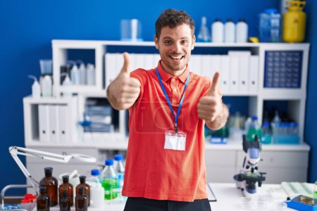 Photo for Young hispanic man working at scientist laboratory approving doing positive gesture with hand, thumbs up smiling and happy for success. winner gesture. - Royalty Free Image