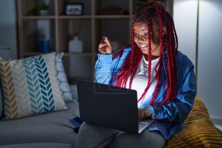Foto de Mujer afroamericana con cabello trenzado usando computadora portátil por la noche con una gran sonrisa en la cara, apuntando con el dedo de la mano hacia el lado mirando a la cámara. - Imagen libre de derechos