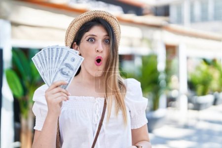 Foto de Young brunette woman holding dollars banknotes scared and amazed with open mouth for surprise, disbelief face - Imagen libre de derechos