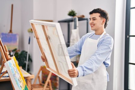 Téléchargez les photos : Jeune homme non binaire artiste souriant confiant tenant dessiner toile au studio d'art - en image libre de droit