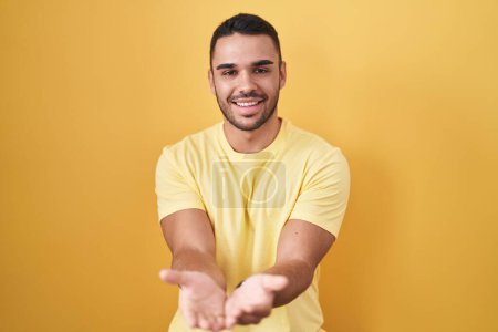 Téléchargez les photos : Jeune homme hispanique debout sur fond jaune souriant avec les mains paumes ensemble recevant ou donnant geste. maintien et protection - en image libre de droit