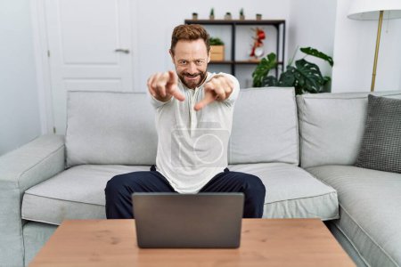 Téléchargez les photos : Homme d'âge moyen en utilisant un ordinateur portable à la maison pointant vers vous et la caméra avec les doigts, souriant positif et joyeux - en image libre de droit