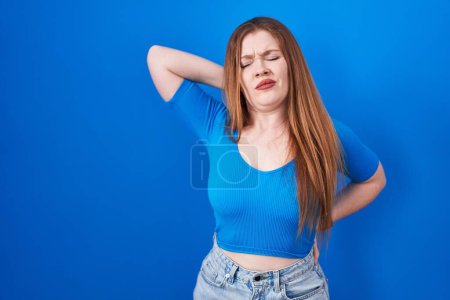 Photo for Redhead woman standing over blue background suffering of neck ache injury, touching neck with hand, muscular pain - Royalty Free Image