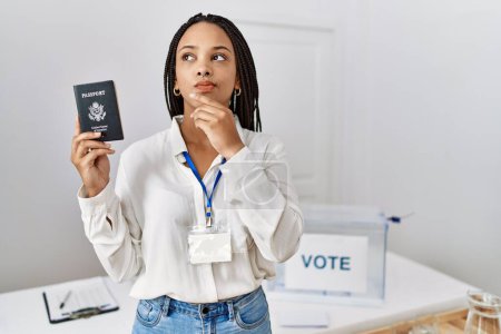 Foto de Mujer afroamericana joven en las elecciones de campaña política celebración de EE.UU. pasaporte cara seria pensando en la pregunta con la mano en la barbilla, reflexivo acerca de la idea confusa - Imagen libre de derechos