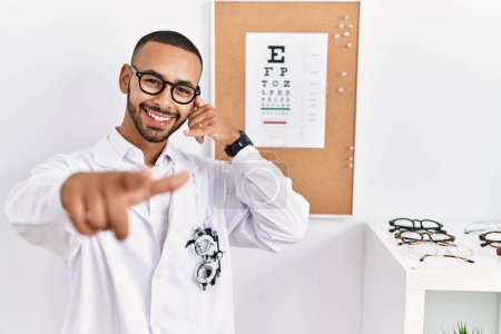 Téléchargez les photos : Un opticien afro-américain debout à côté d'un test de vue souriant parlant au téléphone et vous montrant du doigt. Appelle-moi. - en image libre de droit