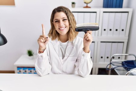 Foto de Mujer hispana joven que trabaja en la clínica de odontología llevando a cabo una prueba de blanqueamiento sonriendo con una idea o pregunta señalando con el dedo con la cara feliz, número uno - Imagen libre de derechos
