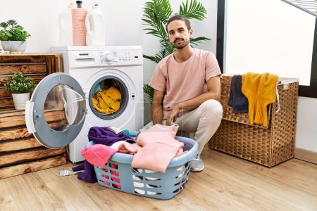 Foto de Young hispanic man putting dirty laundry into washing machine with serious expression on face. simple and natural looking at the camera. - Imagen libre de derechos