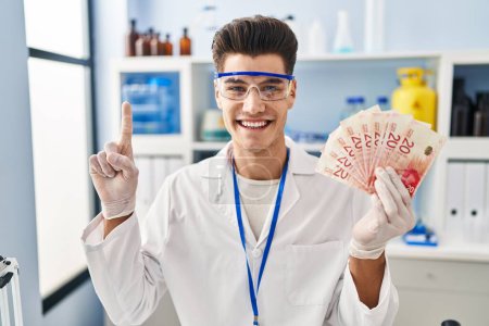 Téléchargez les photos : Jeune homme hispanique travaillant au laboratoire scientifique tenant des shekels souriant avec une idée ou une question pointant du doigt avec un visage heureux, numéro un - en image libre de droit