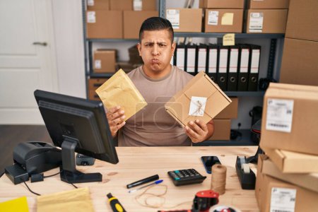 Photo for Hispanic young man working at small business ecommerce holding packages puffing cheeks with funny face. mouth inflated with air, catching air. - Royalty Free Image