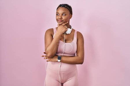Photo for African american woman with braids wearing sportswear and headphones looking confident at the camera smiling with crossed arms and hand raised on chin. thinking positive. - Royalty Free Image