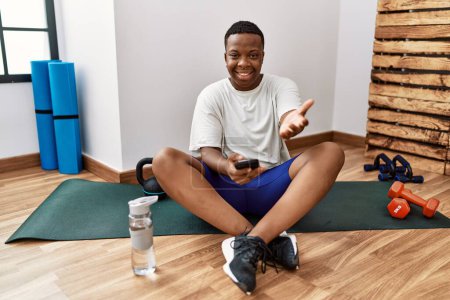Téléchargez les photos : Jeune homme africain assis sur tapis d'entraînement à la salle de gym à l'aide d'un smartphone souriant gai offrant main de paume donnant assistance et acceptation. - en image libre de droit