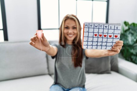 Photo for Young caucasian girl holding period calendar and menstrual cup sitting on the sofa at home. - Royalty Free Image