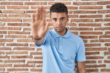 Téléchargez les photos : Jeune homme brésilien debout sur un mur de briques faire arrêter chanter avec la paume de la main. expression d'avertissement avec geste négatif et sérieux sur le visage. - en image libre de droit