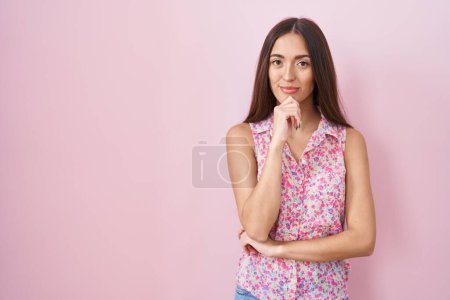 Foto de Mujer hispana joven con el pelo largo de pie sobre fondo rosa mirando con confianza a la cámara con sonrisa con los brazos cruzados y la mano levantada en la barbilla. pensar positivamente. - Imagen libre de derechos