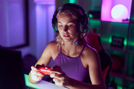 Téléchargez les photos : Jeune femme blonde streamer jouer à un jeu vidéo en utilisant joystick à la salle de jeu - en image libre de droit