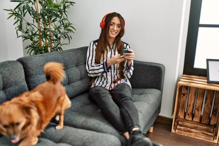 Téléchargez les photos : Jeune femme hispanique utilisant smartphone assis sur le canapé avec chien à la maison - en image libre de droit