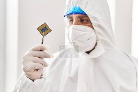 Foto de Joven científico latino usando uniforme de protección covid sosteniendo chip de procesador de CPU en el laboratorio - Imagen libre de derechos