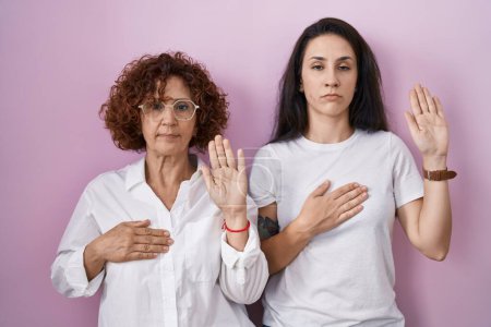 Foto de Madre e hija hispanas usando una camiseta blanca casual sobre fondo rosa jurando con la mano en el pecho y la palma abierta, haciendo un juramento de lealtad - Imagen libre de derechos